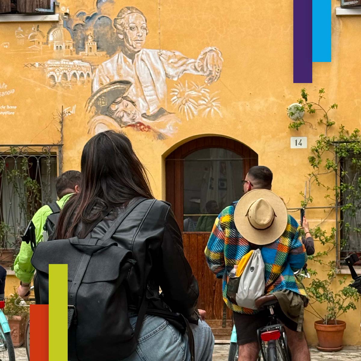 Un gruppo di persone in bicicletta si ferma davanti a una casa con facciata gialla su cui è dipinto un murales con due figure storiche e elementi architettonici. L’atmosfera è curiosa e coinvolta, in un contesto urbano pittoresco.