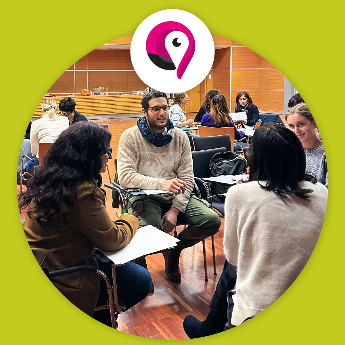 Gruppo di partecipanti seduti in cerchio durante un laboratorio formativo. Alcuni prendono appunti e discutono in coppia, in una sala luminosa con pavimento in legno. In alto è visibile il logo del progetto su sfondo verde.
