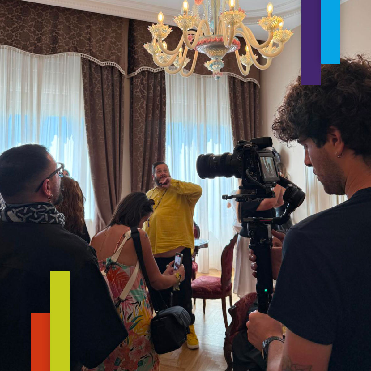 Un gruppo di persone è riunito in una sala elegante con tendaggi pesanti e un grande lampadario di vetro. In primo piano un videomaker riprende la scena con una videocamera montata su stabilizzatore. Tra i presenti, alcuni interagiscono con il videomaker, mentre altri si lasciano riprendere.