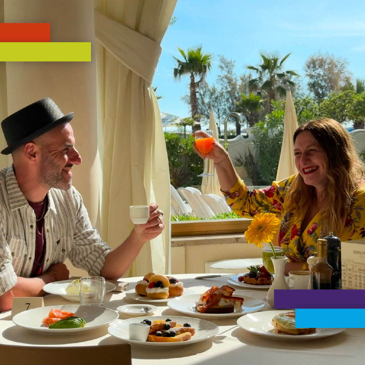 Due persone fanno colazione in un ambiente elegante e soleggiato, con vista su palme e piscina. Ridono e brindano con una spremuta e un caffè, circondati da piatti di dolci e frutta fresca.