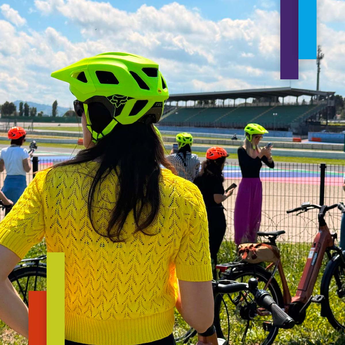 Donna con caschetto da bici giallo fluo osserva un autodromo. Altre persone con caschetti colorati e biciclette sono sparse sul prato vicino alla pista. Atmosfera sportiva e dinamica.