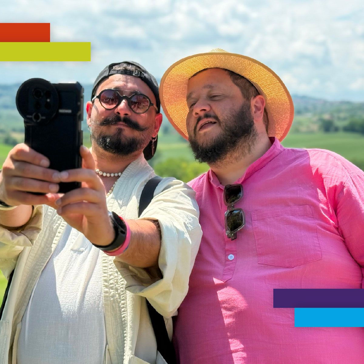 Due uomini si scattano un selfie in un contesto collinare soleggiato. Uno indossa un cappello di paglia e camicia rosa, l’altro un cappellino e collana di perle. Entrambi sorridenti, trasmettono complicità.