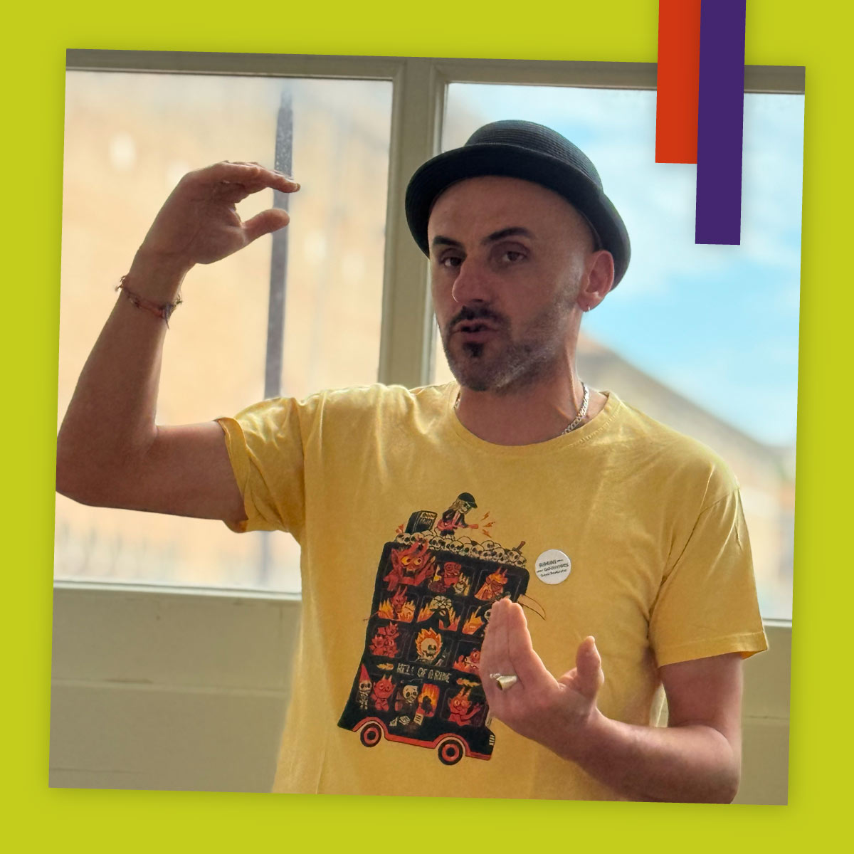 Primo piano di un uomo che parla gesticolando con le mani. Indossa una maglietta gialla con una grafica colorata e un cappello nero. Alle sue spalle si vedono finestre che lasciano entrare molta luce.