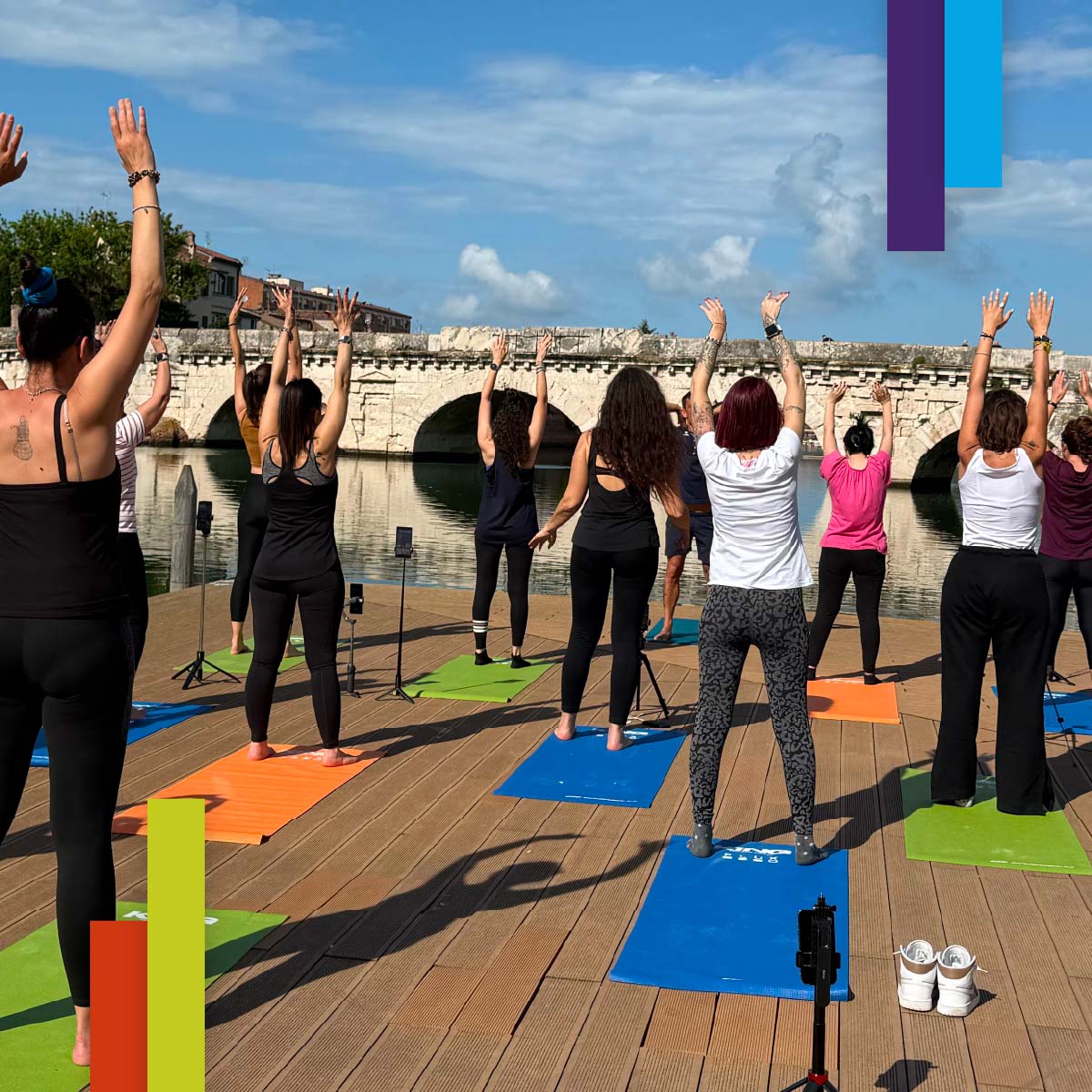 Gruppo di persone su tappetini colorati pratica yoga su una pedana in legno davanti al Ponte di Tiberio a Rimini, con il cielo sereno e l’acqua che riflette il ponte. Un momento di benessere e connessione con l’ambiente.