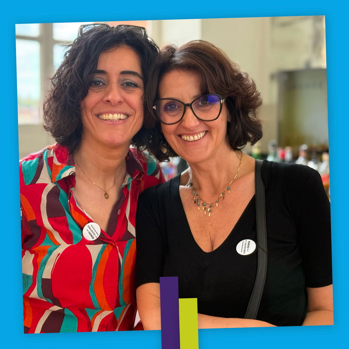 Due donne sorridono abbracciate davanti a un tavolo da buffet. Una indossa una camicia colorata, l’altra una maglia nera con occhiali. Entrambe portano un badge rotondo e trasmettono un senso di complicità e gioia.