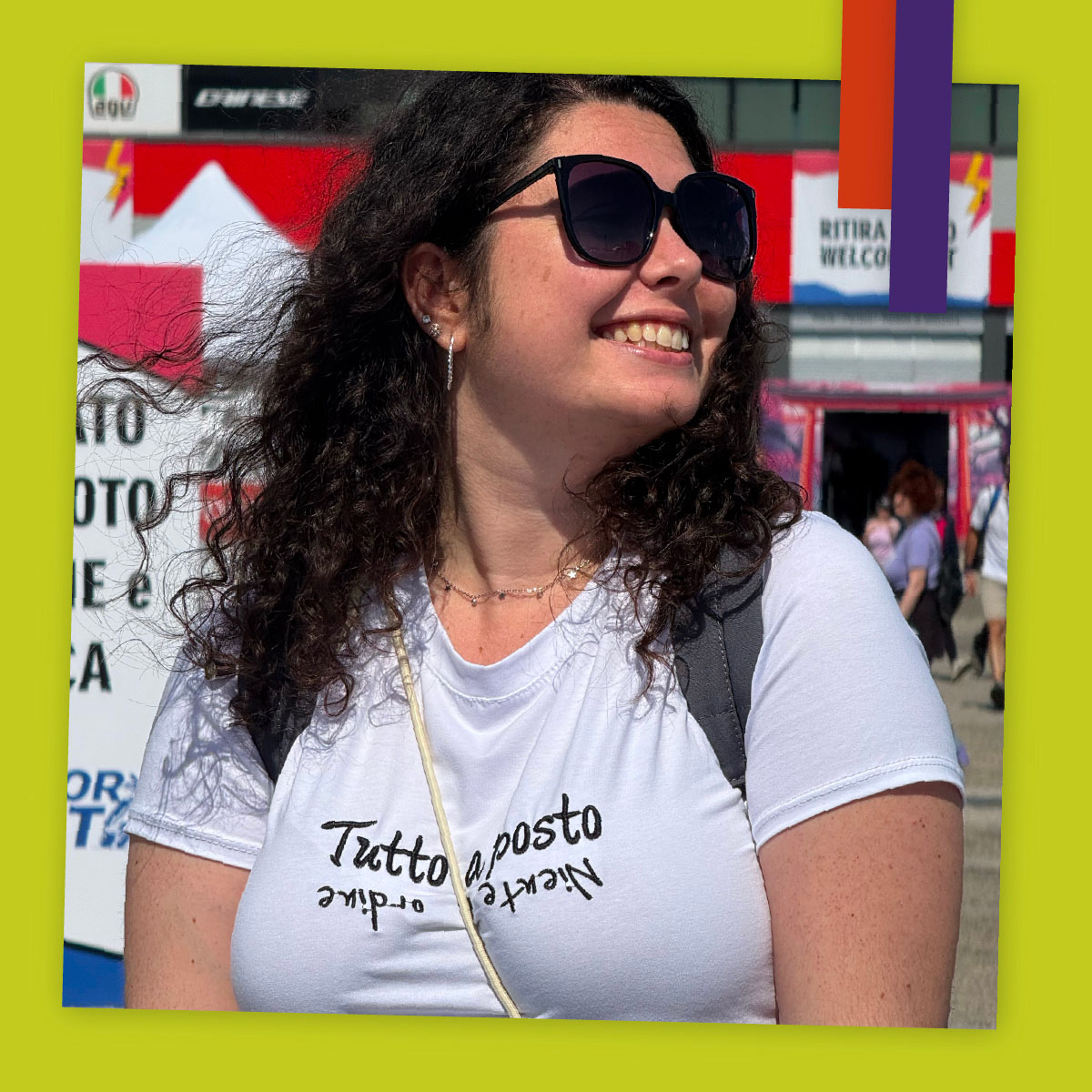 Donna sorride in primo piano con una maglietta con scritta “Tutto apposto – Niente in ordine”, sullo sfondo un evento all’aperto.