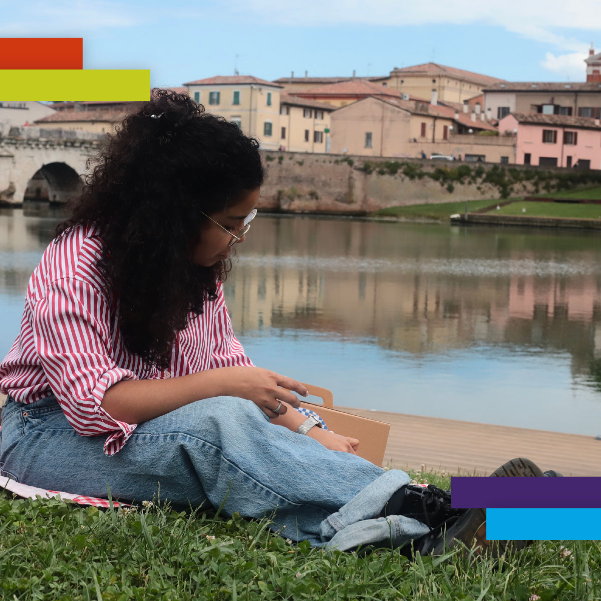 Una giovane donna dai capelli ricci siede sull’erba vicino al fiume, intenta a disegnare o scrivere su un blocco appoggiato alle ginocchia. Alle sue spalle si vede il borgo antico di Rimini e il ponte di Tiberio.