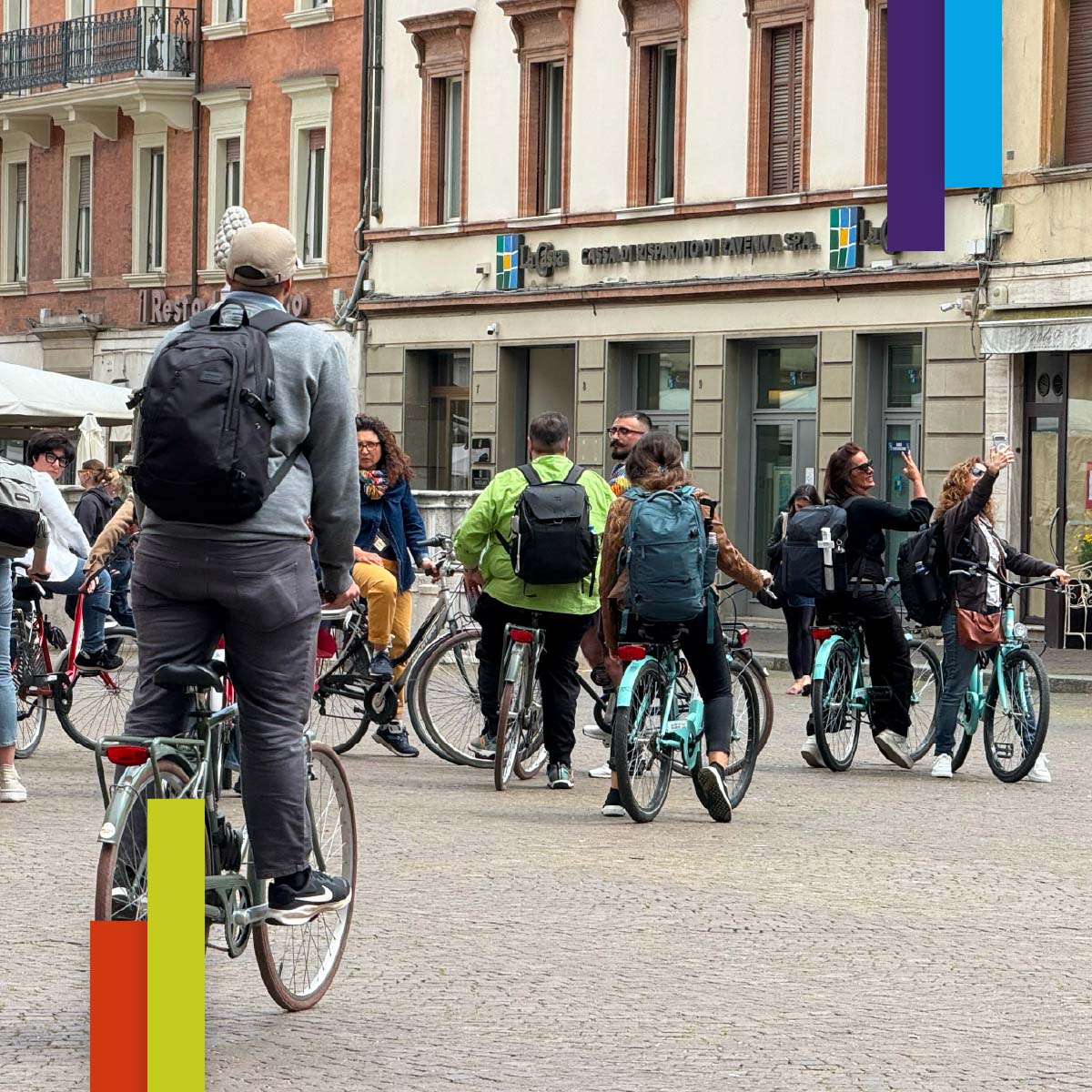 Un gruppo numeroso di persone in bicicletta attraversa una piazza del centro storico di Rimini, circondata da edifici in stile tradizionale. Alcuni ciclisti scattano foto, creando un’atmosfera dinamica e turistica.