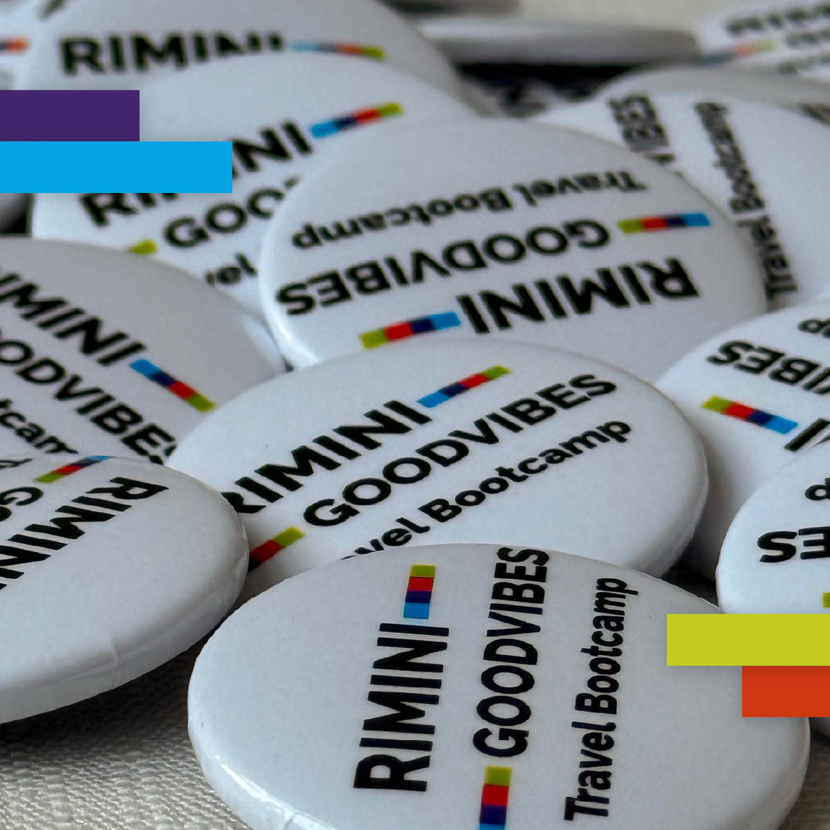 Dettaglio ravvicinato di una serie di spille promozionali bianche con il logo “Rimini Good Vibes Travel Bootcamp”, sparse su una superficie chiara.
