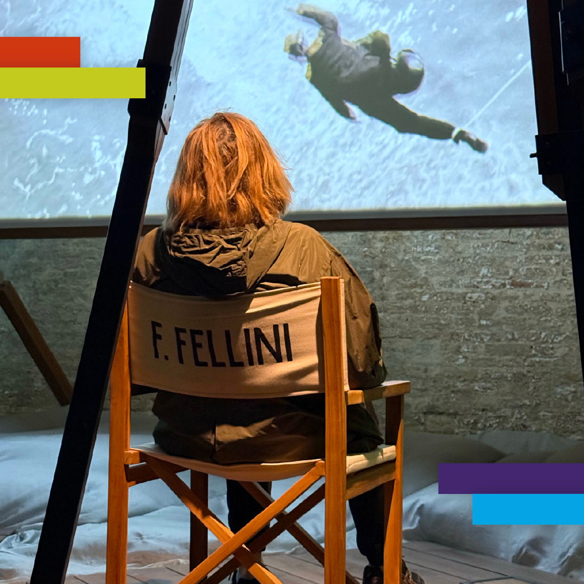 Una persona di spalle è seduta su una sedia da regista con la scritta "F. Fellini", guardando una proiezione cinematografica su uno schermo. L’ambiente è interno, con luce soffusa, evocando un omaggio al grande regista riminese.