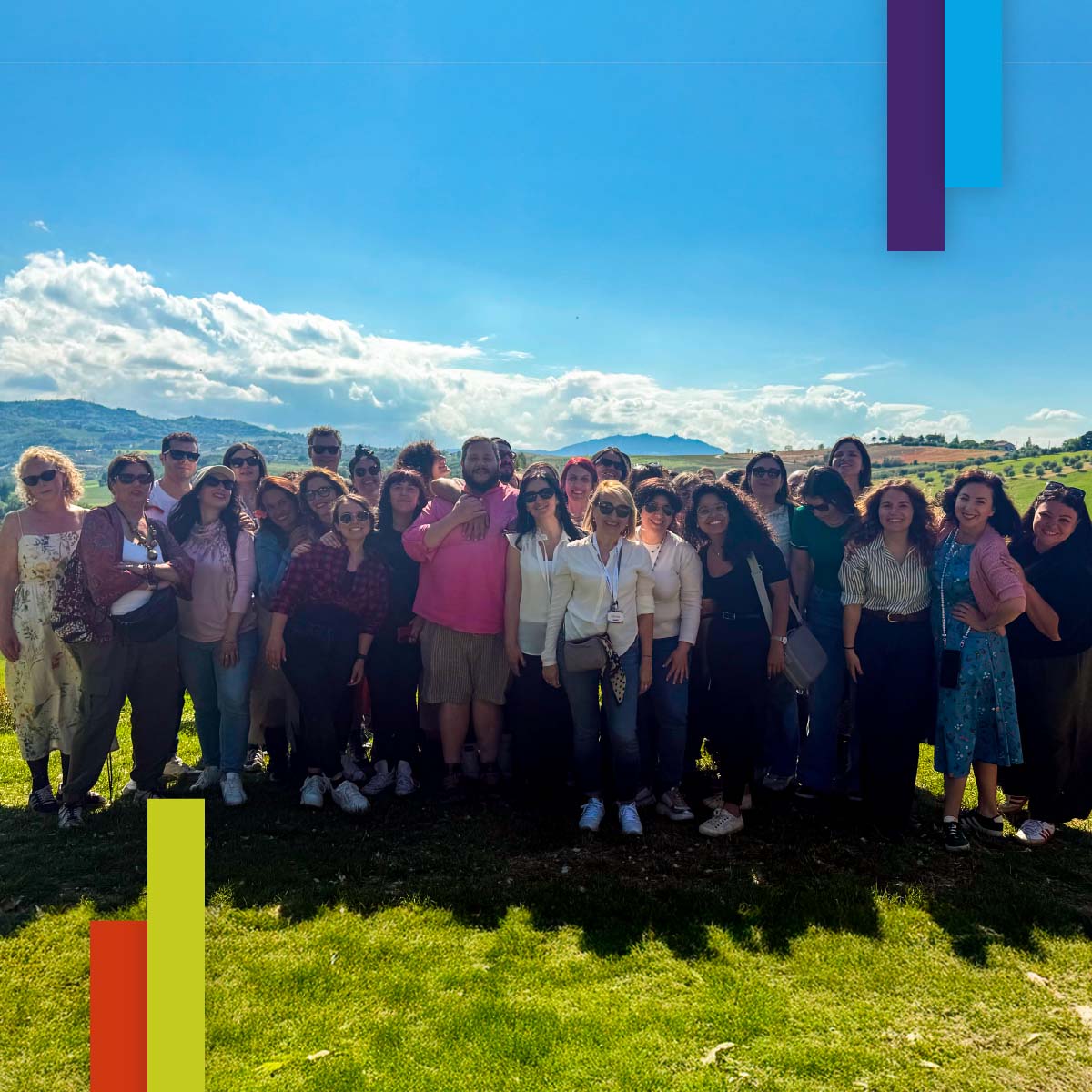 Foto di gruppo all’aperto di circa trenta persone sorridenti, in posa su un prato verde con colline e cielo azzurro sullo sfondo. L’immagine trasmette un forte senso di comunità, inclusività e condivisione.