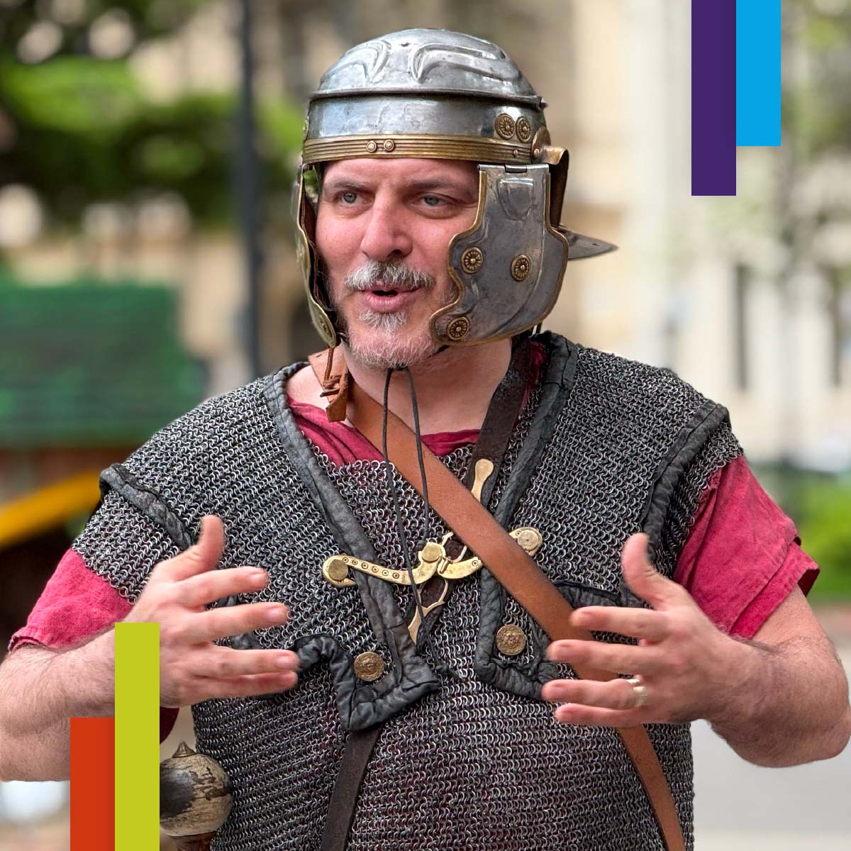 Uomo in costume da legionario romano con elmo e armatura, mentre parla gesticolando. Lo sfondo è sfocato, probabilmente urbano. L’immagine evoca una rievocazione storica o visita guidata.