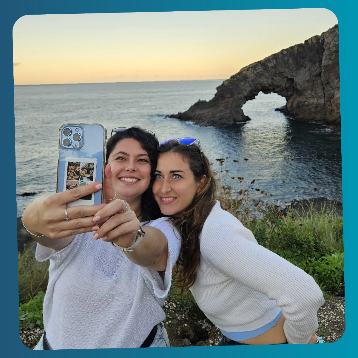 Due giovani donne sorridono scattando un selfie con il mare di Pantelleria e l’arco naturale dell’Arco dell’Elefante sullo sfondo al tramonto.