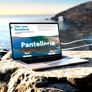 2 Laptop poggiato su una roccia con vista sul mare e sull’Arco dell’Elefante di Pantelleria. Sullo schermo compare la homepage del sito Visit Pantelleria Island, con il titolo “Ciao, sono Pantelleria. Come posso rendere magica la tua esperienza?” e una grande immagine aerea dell’isola.