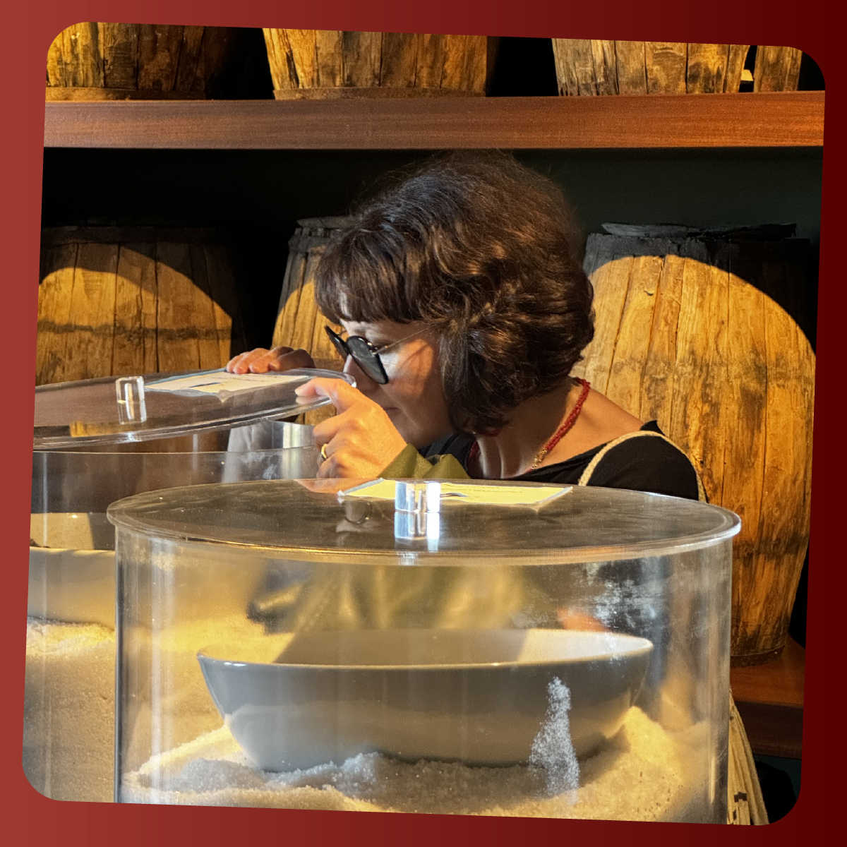 Donna con occhiali osserva e annusa il contenuto di un contenitore trasparente riempito di sale, in un ambiente con botti di legno sullo sfondo. L’immagine rappresenta un’esperienza sensoriale legata ai profumi e ai prodotti dell’isola.