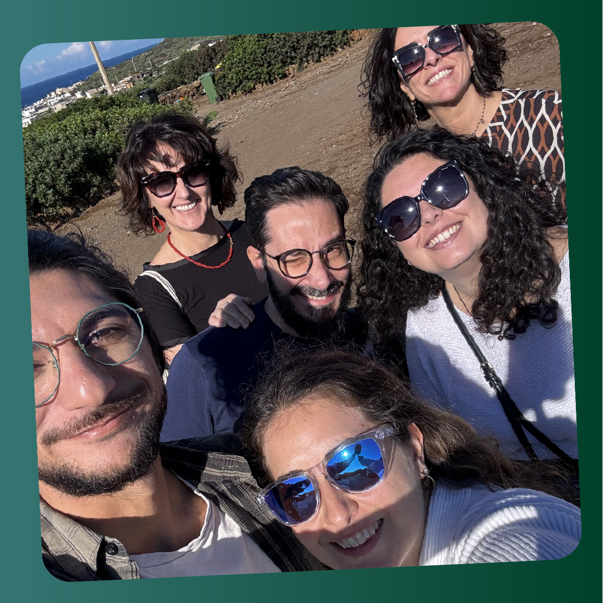 Gruppo di sei persone sorride all’aperto durante una giornata di sole a Pantelleria, con il mare e la vegetazione dell’isola sullo sfondo.
