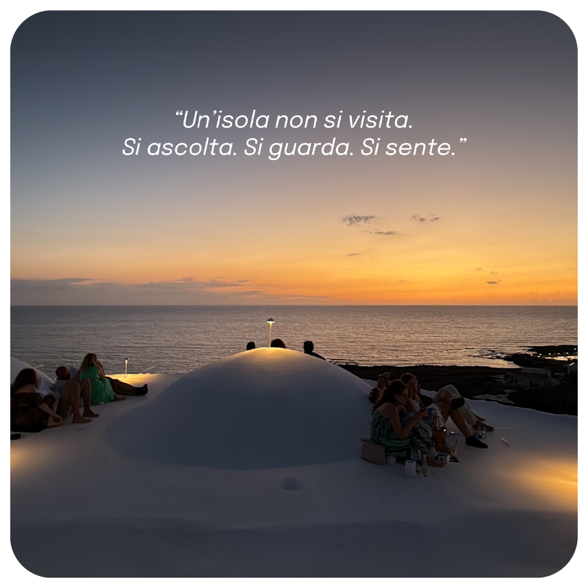 “Tramonto sul mare di Pantelleria con persone sedute sui tetti bianchi dei dammusi. Il cielo è color arancio e rosa. In alto la frase: ‘Un’isola non si visita. Si ascolta. Si guarda. Si sente.’”