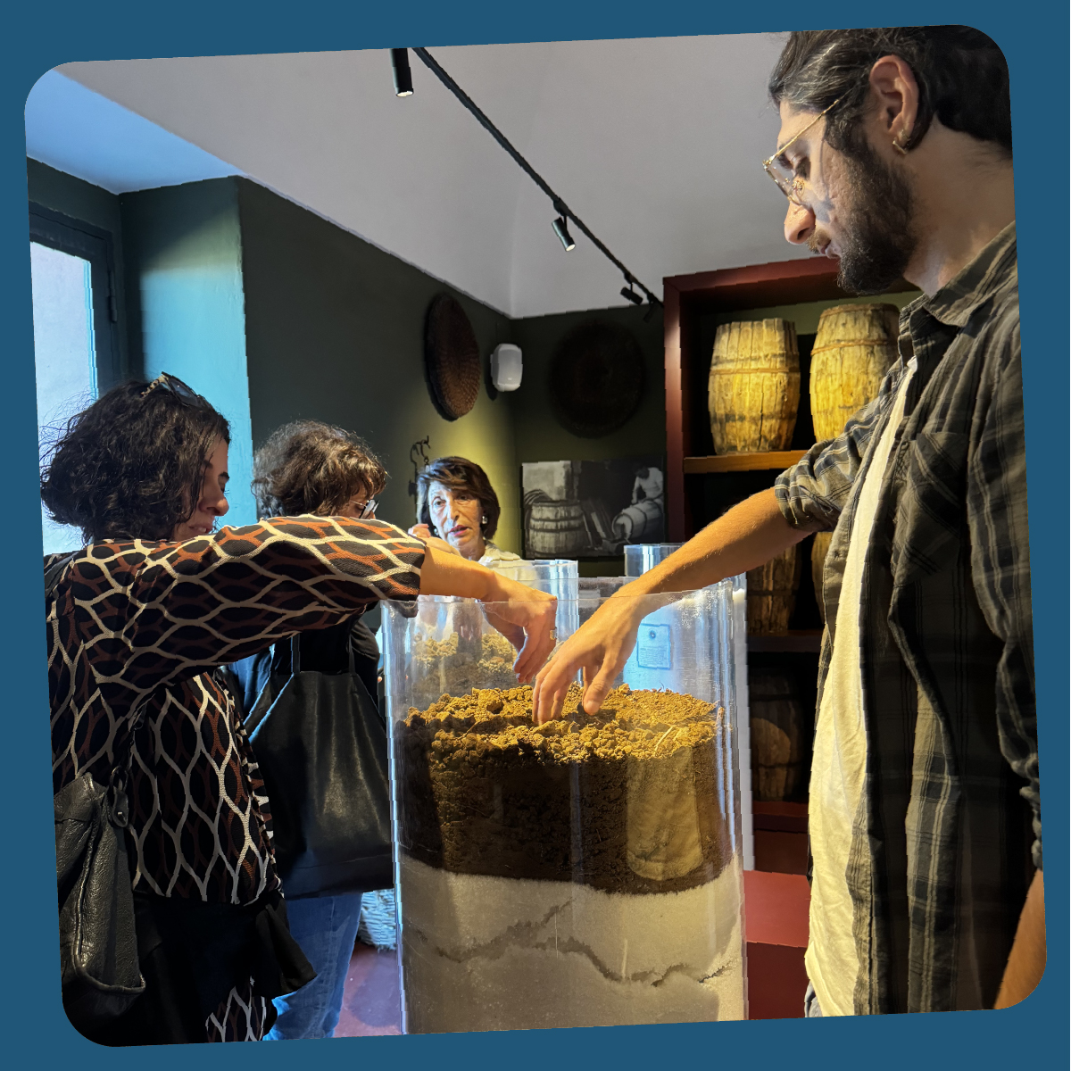 Gruppo di persone all’interno di un museo o laboratorio didattico, intente a toccare campioni di terra e materiali naturali esposti in cilindri trasparenti. L’attività mostra un momento esperienziale di conoscenza del territorio di Pantelleria attraverso i suoi elementi naturali.