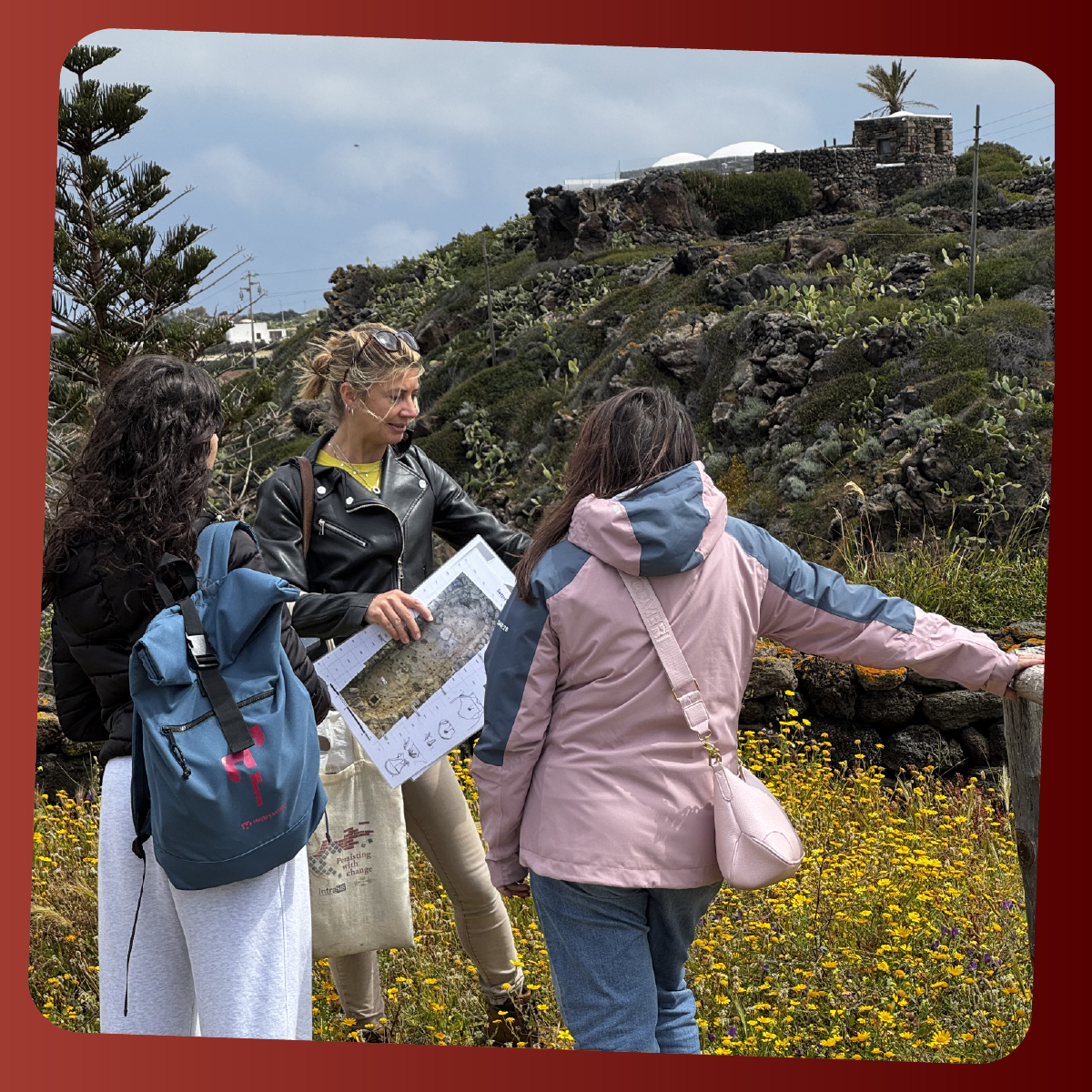 Guida ambientale illustra una mappa a due partecipanti durante un’escursione tra i fiori e i muretti di pietra lavica dell’isola di Pantelleria. Il gruppo ascolta le spiegazioni sul paesaggio e sulla storia del luogo.