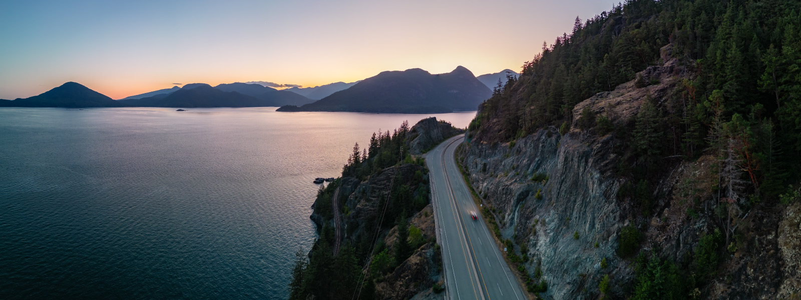 Strada panoramica costiera al tramonto tra mare e montagne, simbolo del travel trend 2026 legato al road trip lento e al viaggio on the road.